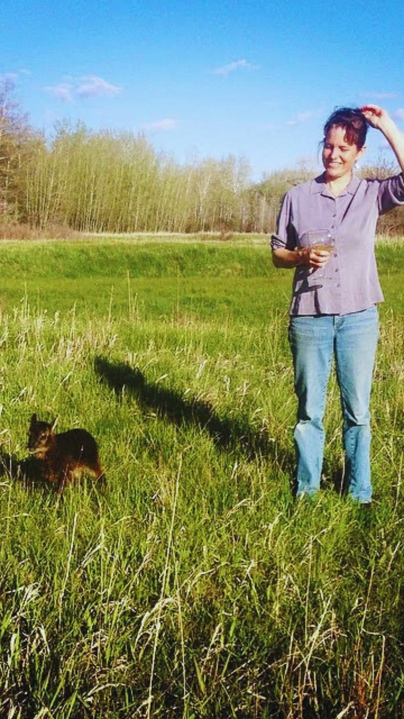 Lorca standing in long green grass with a bird lifted in one hand, a dog at her feet