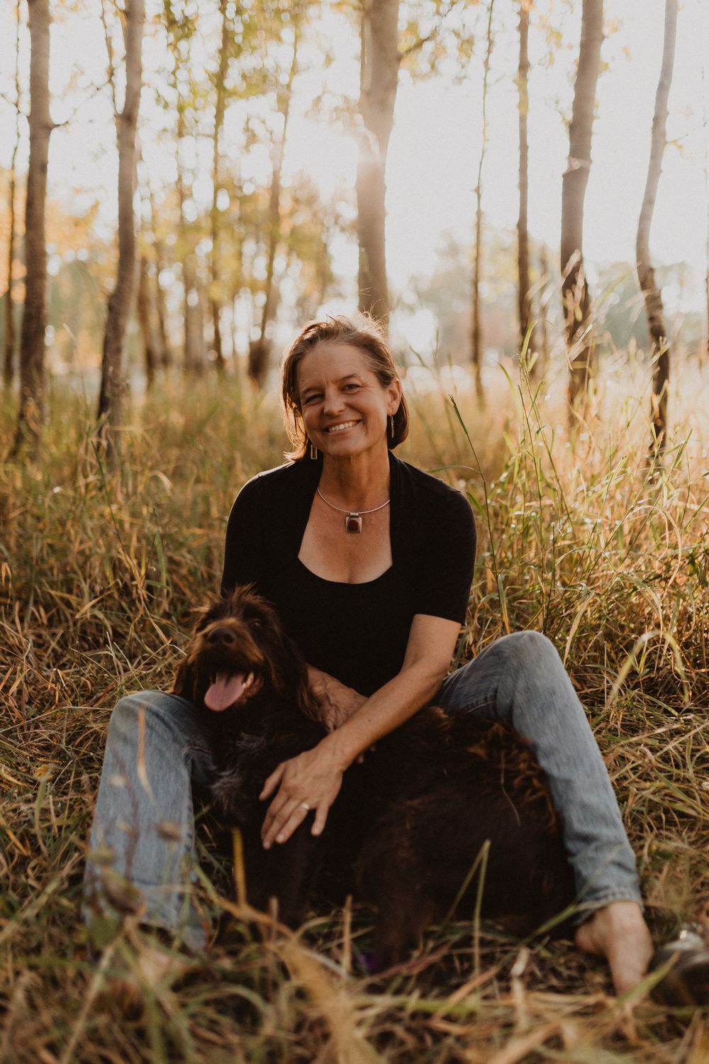 Lorca sitting barefoot in tall gold grass among aspens, laughing, with a large dark curly-coated dog leaning into her lap