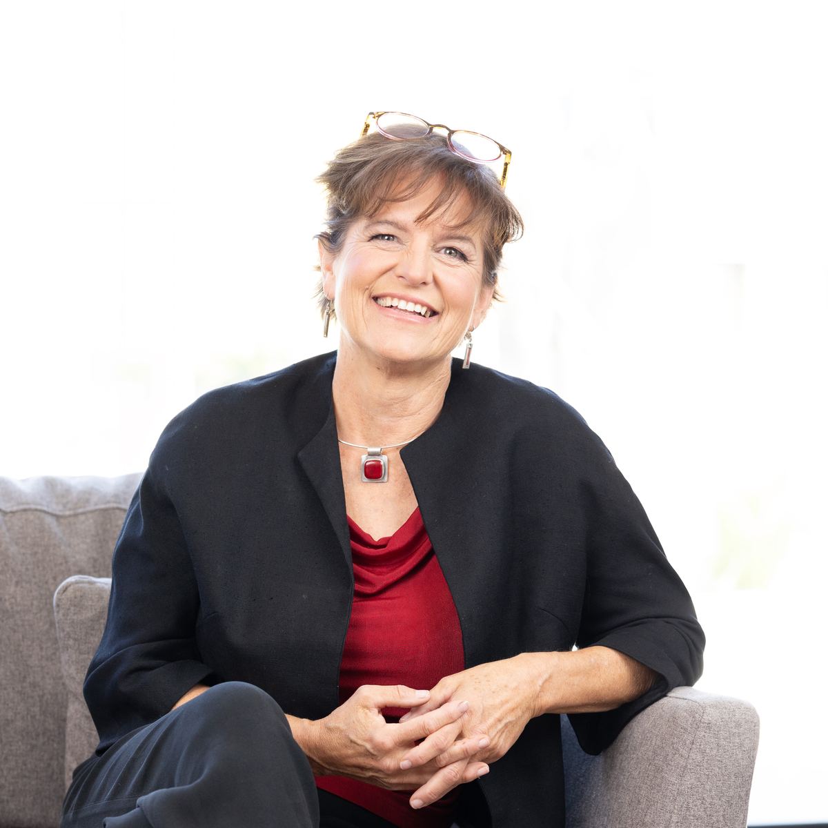 Lorca sitting on a sofa in a black jacket over a red top, laughing, glasses pushed up on her head