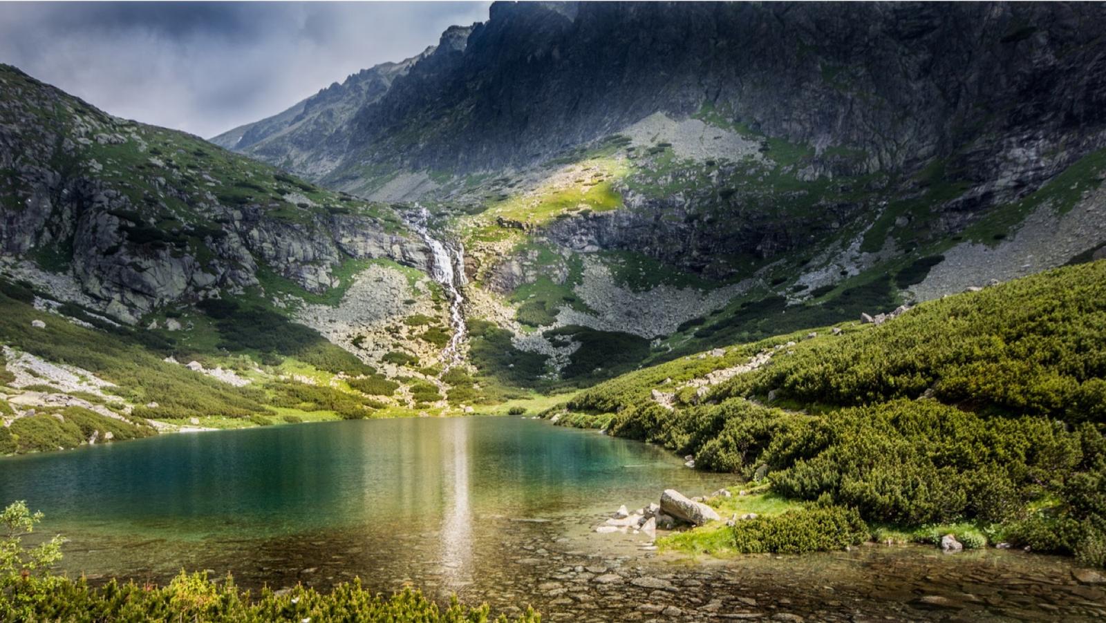 A clear mountain tarn beneath dark peaks, a waterfall threading down to the water