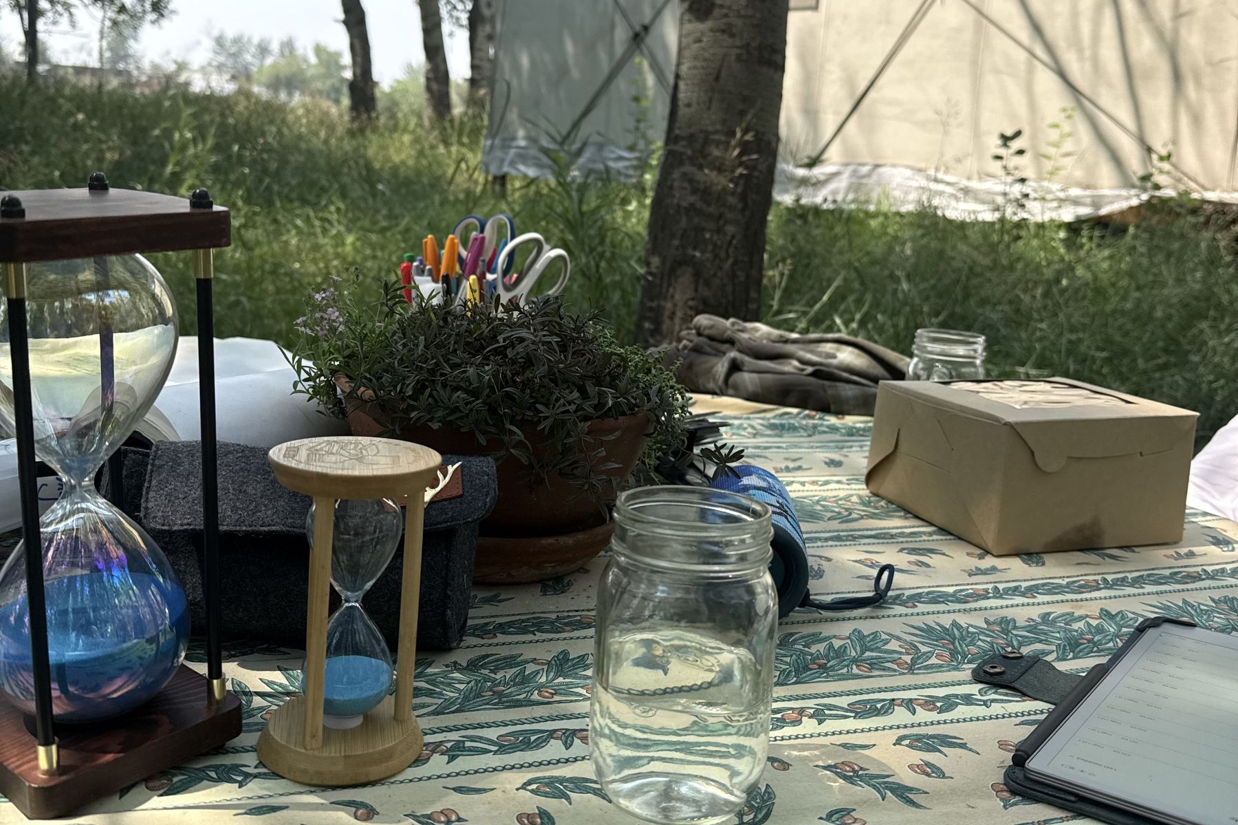 A table set outside the wall tent — two hourglasses, a pot of thyme, a jar of coloured pens and scissors, work gloves and a mason jar of water on a block-printed cloth